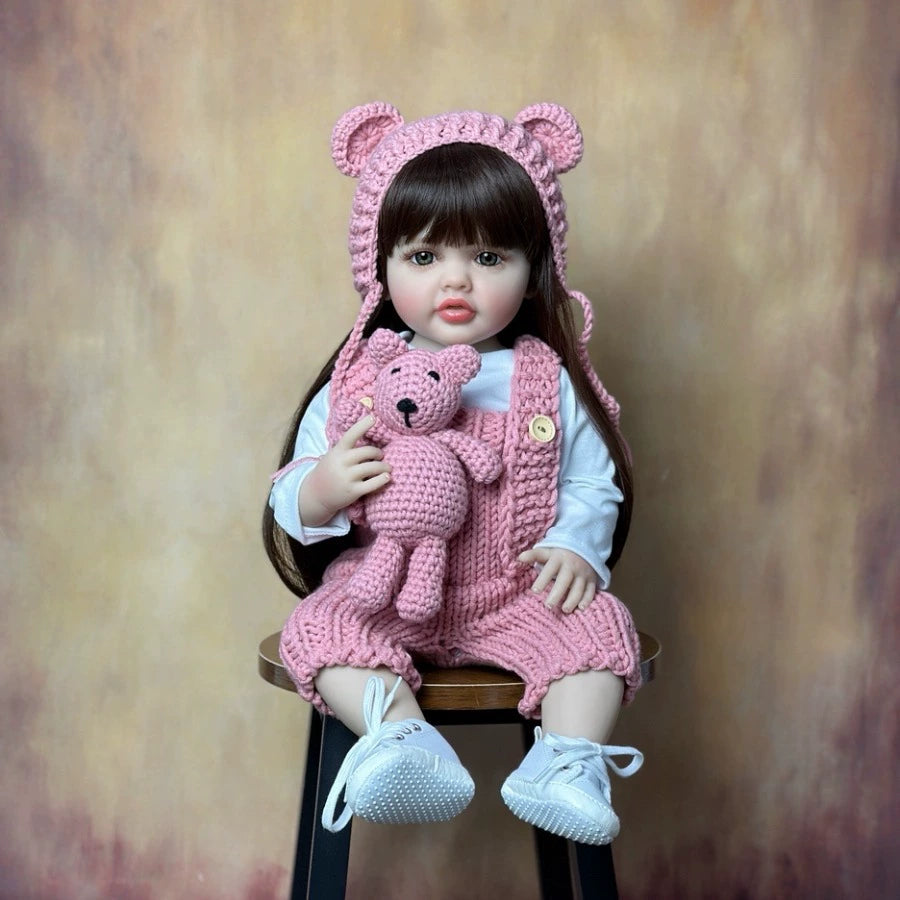 Doll wearing a pink bear hat and outfit, holding a pink teddy bear, sitting on a wooden stool against a brown background.